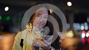 happy female city dweller with mobile phone in night city street, sending message by smartphone