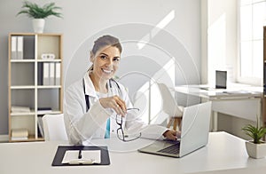 Happy female doctor sitting at working desk with laptop, looking at camera and smiling