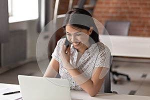 Happy female employee speak on cellphone working on laptop