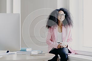 Happy female entrepreneur has bushy Afro hair, wears rosy jacket and black trousers, holds cup of drink, poses at windowsill in