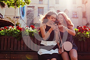 Happy female friends checking social network using phone by cafe on city street. Summer leisure