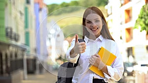 Happy female high-schooler with rucksack and books gesturing thumbs-up, student