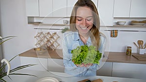 Happy female housewife takes bunch of salad leaves in hands and poses with smile at camera, standing in middle of