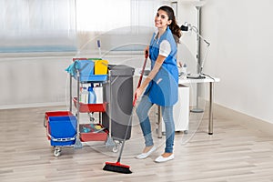 Happy Female Janitor Cleaning Floor With Broom In Office