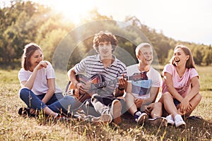 Happy female and male students enjoy picnic outdoor, sit grouped together, laugh and joke among themselves, sing songs to guitar,