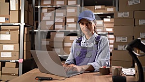 Happy female manager working on laptop in warehouse, preparing parcels for delivery. Storehouse worker smile for camera