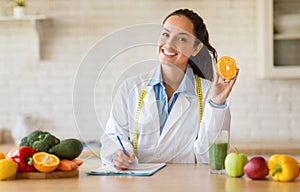 Happy female nutrition adviser holding orange fruit, making meal plan for client and smiling at camera