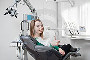 Happy female patient with braces on teeth sitting in dental chair, smiling and showing thumbs up after treatment