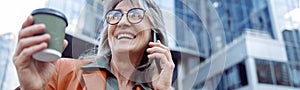 Happy female pensioner with cup of drink n talks on mobile phone on modern city street