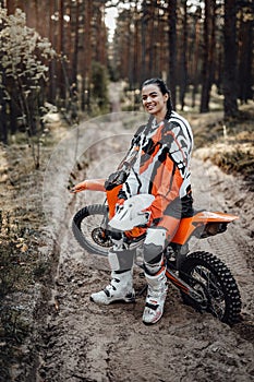 Happy female racer wearing motorcycle outfit sitting on her bike off-road in the woods