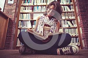 Happy female student against bookshelf reading a book on the library floor