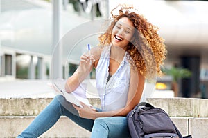 Happy female student sitting outside with book and pen