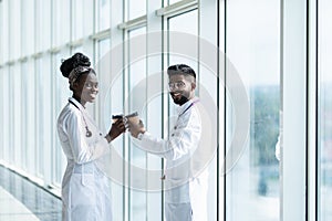 Happy african female surgeon holding coffee mug while looking at indian coworker in hospital