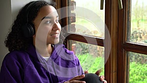 Happy female teenager young biracial African American woman teen sitting by a window listening to music on wireless headphones