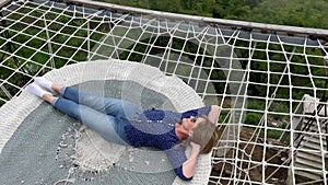 A happy female tourist lying on a hammock couch above green abyss in the mountains. Woman stretching and relaxing in