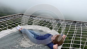 Happy female traveler lying on hammock net couch in the mountains above wild forest covered by white mist. Misty magical