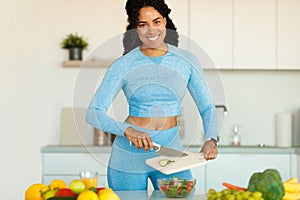 Happy fit black woman in sportswear preparing fresh vegetable salad, standing in the kitchen after domestic workout