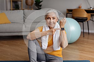 Happy fit senior woman pointing at activity fitness tracker and smiling at camera, having break in her training at home