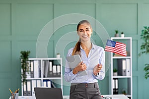 Happy foreign language teacher with USA flag standing near workdesk and holding digital tablet, smiling at camera