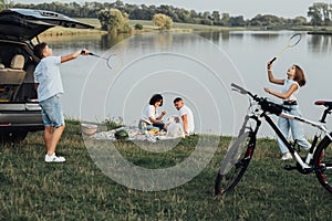 Happy Four Members Family with Pet Dog Having Picnic Outdoors, Two Teenage Children Playing Badminton While Parents