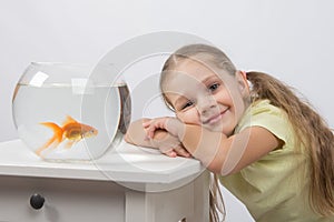 Happy four-year girl put her head in his hands sitting in front of a goldfish