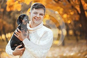 Happy free time with beloved dog! Handsome young man staying in autumn park smiling and holding cute puppy dachshund.