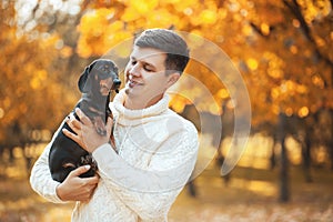 Happy free time with beloved dog! Handsome young man staying in autumn sunny park smiling and holding cute puppy dachshund
