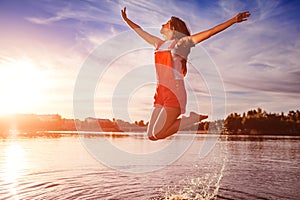 Happy and free young woman jumping and raising arms on river bank. Freedom. Active lifestyle