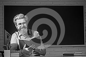 Happy friendly young teacher or post graduate male student standing holding book in front of a blank chalkboard.