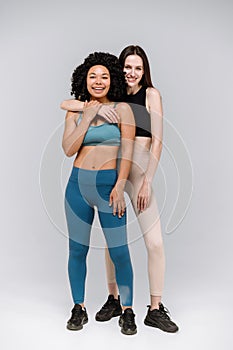 Happy friends in activewear standing together in gym isolated on gray background