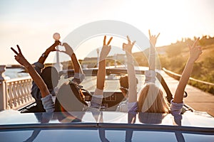 Happy friends in cabriolet with raised hands driving on sunset