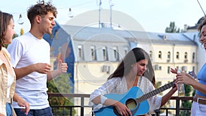 Happy friends dancing at a party while lady playing the guitar, enjoying music on the rooftop outdoors. Lifestyle