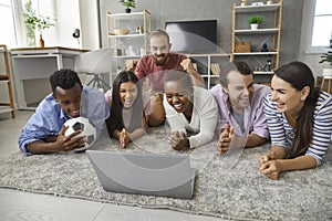 Happy friends diverse group watching football match on laptop lying on floor