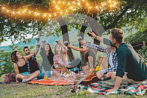 Happy friends having fun at picnic dinner with vintage lights outdoor next vineyard - Young people cheering with red wine on