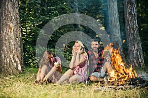 Happy friends having fun at picnic in woods. Bearded man with his girlfriend and her sister sitting near campfire. Blond