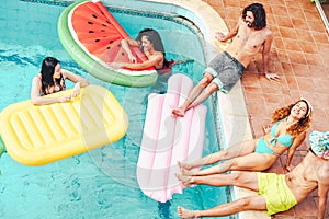 Happy friends having fun in swimming pool during summer tropical vacation - Young people relaxing and floating on air lilos