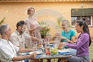 Happy friends laughing with big smile around the table in a summer diner. Woman serves the food at house patio. Smiles