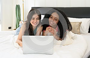 Happy friends using laptop together at home. Two smiling women lying on front and watching media on notebook in a bed
