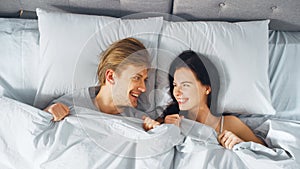 Happy and Fun Young Couple Lying under Blanket on a Bed, Looking at Each other and Smiling. Lovely