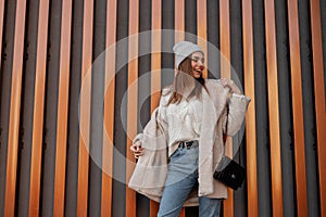 Happy fun young woman in a fashionable hat in a knitted vintage sweater in blue jeans in a stylish warm eco fur coat with a trendy