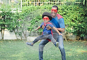 Happy funny African American father carrying little boy flying in superhero costume in garden at home. Happy loving Black African