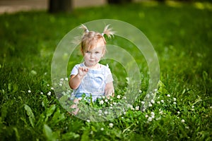 Happy funny blonde baby girl with two little braids in white t-shirt and jeans bodykit sitting on green grass background