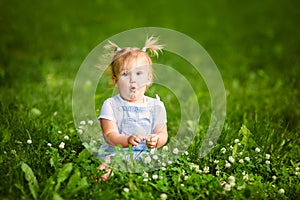 Happy funny blonde baby girl with two little braids in white t-shirt and jeans bodykit sitting on green grass background