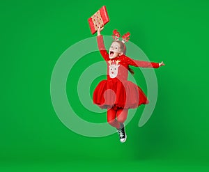 Happy funny child girl in red Christmas reindeer costume jumping  with gift on greeen   background