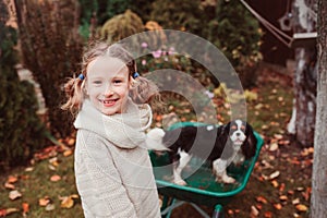 Happy funny child girl riding her dog in wheelbarrow in autumn garden, candid outdoor capture