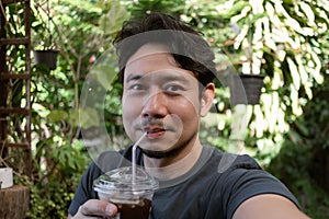 Happy funny face Asian man selfie himself drinks fresh iced coffee in the cafe.