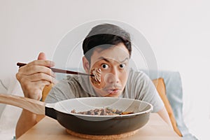 Happy funny face of man eating homemade food in the pan with chopsticks.
