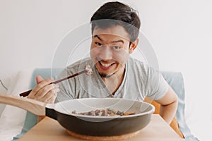 Happy funny face of man eating homemade food in the pan with chopsticks.