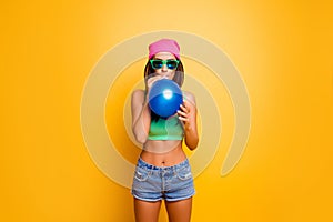 Happy funny hipster girl in pink hat inflate balloon near yellow wall