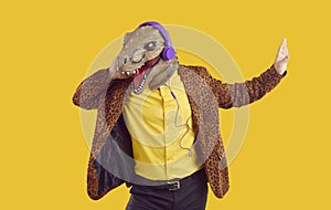 Happy funny man in dinosaur mask and headphones listening to music on yellow studio background